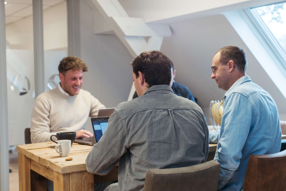 Drie collega&rsquo;s in overleg aan een houten tafel, laptops open en koffie binnen handbereik. Samenwerken aan online succes in een relaxte, professionele sfeer.