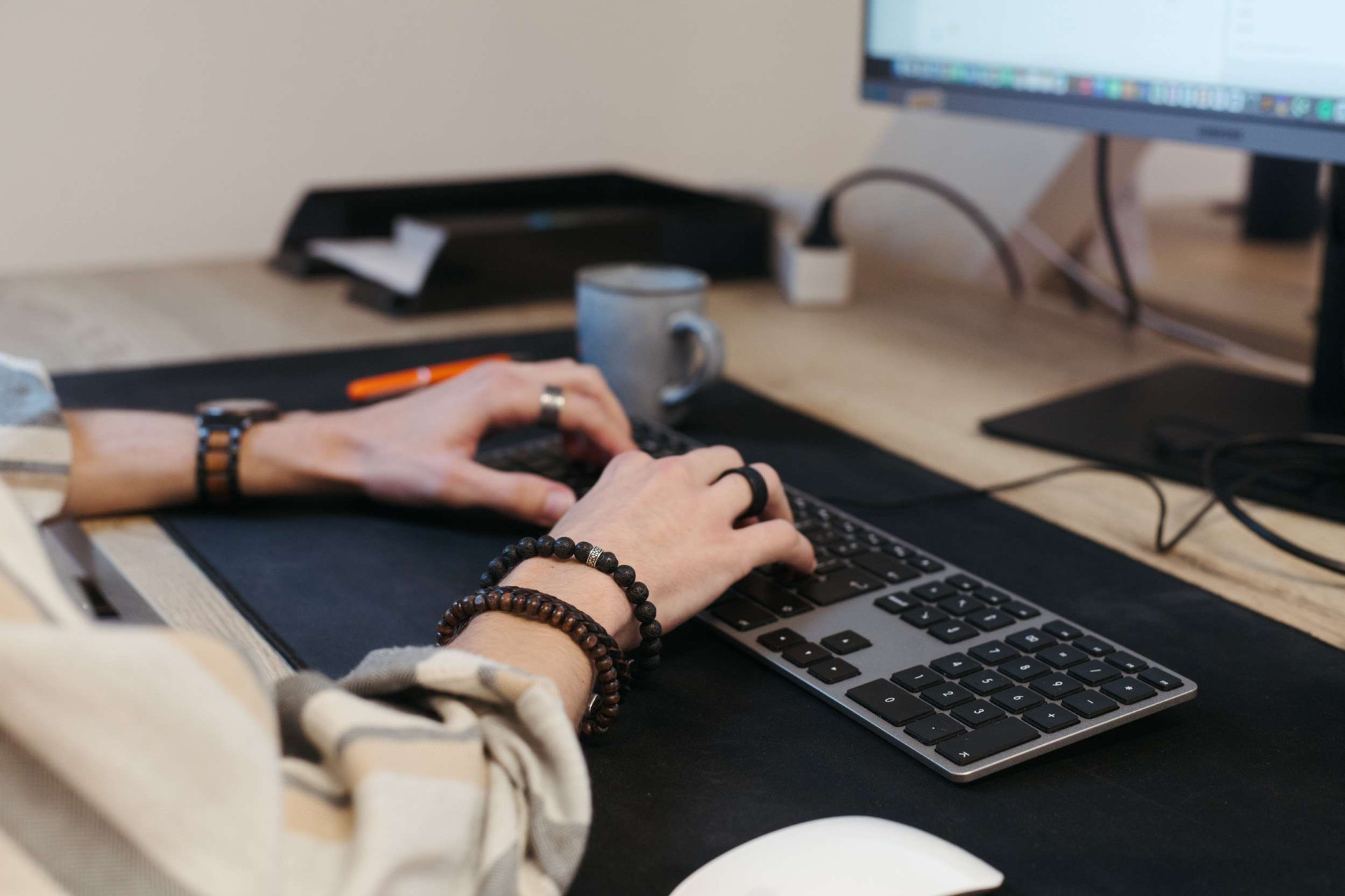 Close-up van handen op het toetsenbord met armbanden om de pols en een koffiemok op de achtergrond. Volle focus op het scherm tijdens een productieve werkdag.