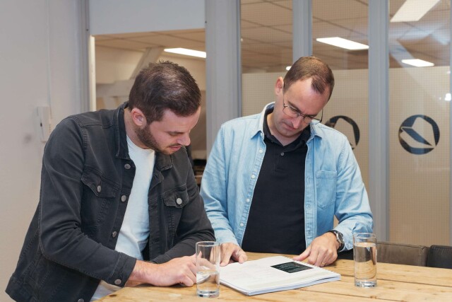 Twee mannen staan aan een tafel en bekijken samen een document, terwijl ze iets aanwijzen en bespreken. Op de tafel staan twee glazen water.