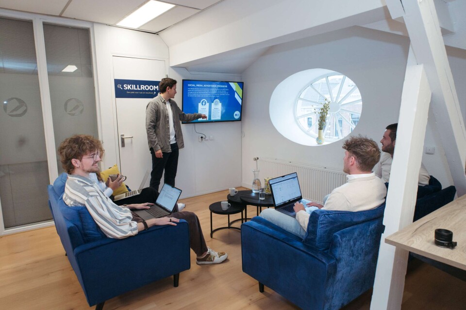 Vergadering tussen online marketeers waarvan er iemand staat te presenteren en een aantal op een blauwe bank zitten